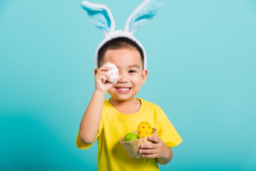 Asian cute little child boy smile beaming wearing bunny ears and a yellow T-shirt, standing to holds colored easter eggs instead of eyes on blue background with copy space for text