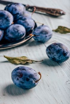 Ripe plums on metal plate and light wooden surface with leaves