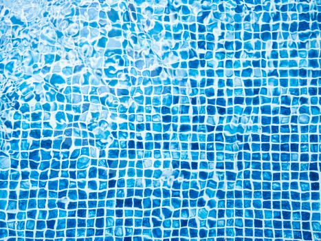 The light reflected on the top of the water surface of the pool lined with blue mosaic tiles below. Water ripples on blue-tiled swimming pool background.