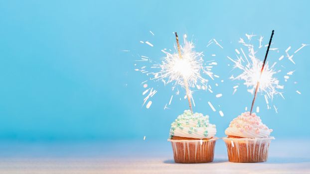 Two cupcakes decorated with colorful sprinkles and sparklers. Festive cupcakes on blue background with copy space left for text or design. Banner