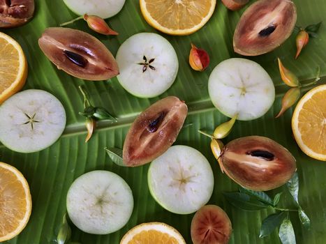 Background of fresh tropical exotic fruits on the banana leaf. Top view. Fruits sliced sapodilla, apple and orange.
