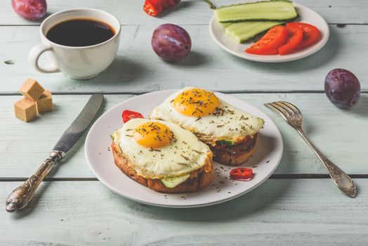 Sandwiches with vegetables and fried egg on white plate, cup of coffee and some fruits over wooden background.