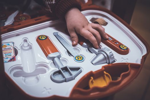 A kid is going to play with a hammer, tweezers, stethoscope, syringe and other toy tools from a toy medical briefcase