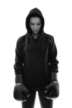 Black and white photo of a focused young beautiful female boxer with hoodie and boxing gloves looking down, backlit silhouette studio shot isolated on white background. Focus, calm, sport concept.