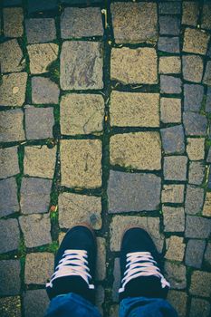 Top down view of feet in a pair of skater sneakers on a cobblestone pavement. Cobblestone patterned background. Minimalist urban style. Feet on cobblestones.