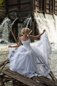 Young blonde woman in a white wedding dress near the waterfall