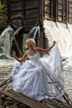 Young blonde woman in a white wedding dress near the waterfall