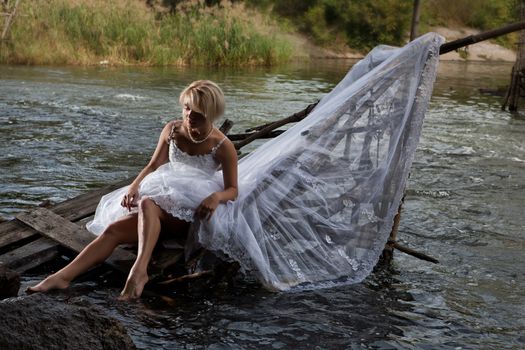 Young blonde woman in a white wedding dress near the waterfall