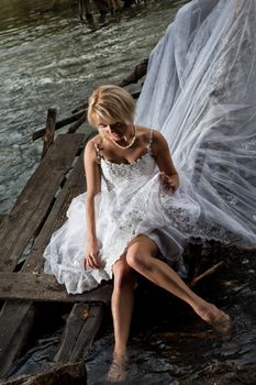 Young blonde woman in a white wedding dress near the waterfall