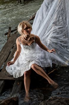 Young blonde woman in a white wedding dress near the waterfall