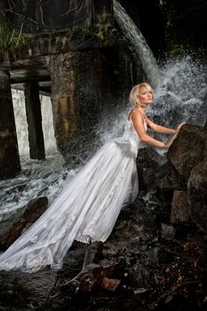 Young blonde woman in a white wedding dress near the waterfall