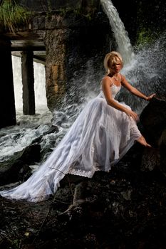 Young blonde woman in a white wedding dress near the waterfall