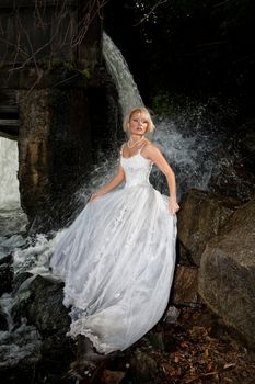 Young blonde woman in a white wedding dress near the waterfall