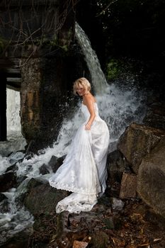 Young blonde woman in a white wedding dress near the waterfall