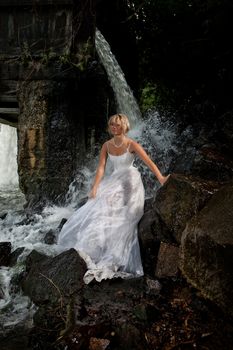 Young blonde woman in a white wedding dress near the waterfall