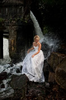 Young blonde woman in a white wedding dress near the waterfall