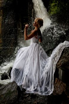 Young blonde woman in a white wedding dress near the waterfall