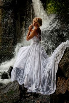 Young blonde woman in a white wedding dress near the waterfall