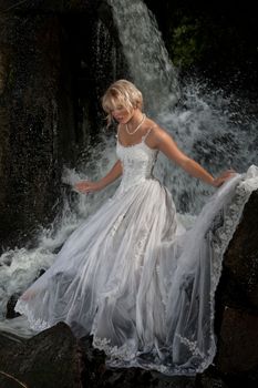Young blonde woman in a white wedding dress near the waterfall