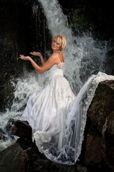 Young blonde woman in a white wedding dress near the waterfall