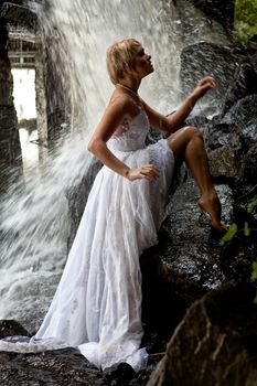 Young blonde woman in a white wedding dress near the waterfall