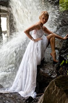 Young blonde woman in a white wedding dress near the waterfall