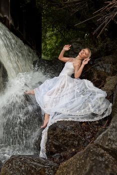 Young blonde woman in a white wedding dress near the waterfall