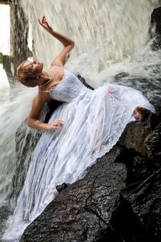 Young blonde woman in a white wedding dress near the waterfall