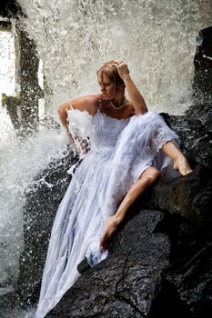 Young blonde woman in a white wedding dress near the waterfall