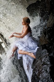 Young blonde woman in a white wedding dress near the waterfall