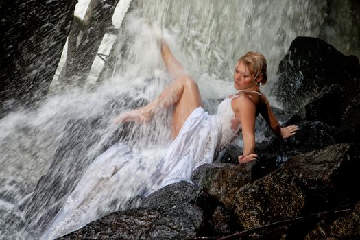 Young blonde woman in a white wedding dress near the waterfall