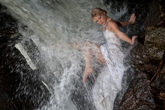 Young blonde woman in a white wedding dress near the waterfall