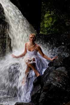 Young blonde woman in a white wedding dress near the waterfall