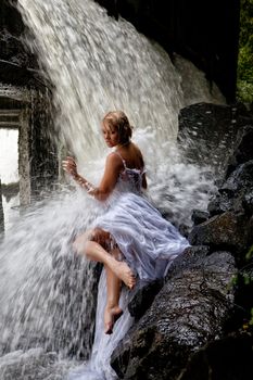 Young blonde woman in a white wedding dress near the waterfall