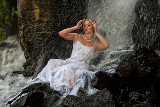 Young blonde woman in a white wedding dress near the waterfall