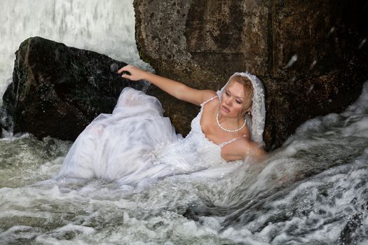 Young blonde woman in a white wedding dress near the waterfall