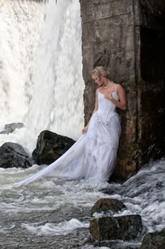 Young blonde woman in a white wedding dress near the waterfall