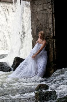 Young blonde woman in a white wedding dress near the waterfall