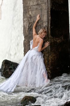 Young blonde woman in a white wedding dress near the waterfall