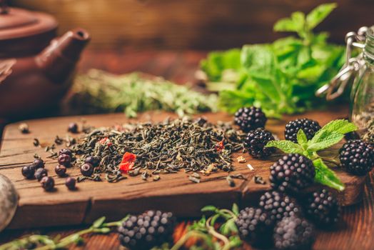 Heap of Dry Green Tea and Fresh Blackberries on Wooden Cutting Board. Bundles of Mint and Thyme Leaves. Clay Tea Pot.