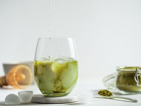 Iced Matcha Latte Tea with pouring milk drops into tumbler glass. Matcha latte and ingredients on white marble background. Copy space for txt or design