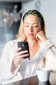 Beautiful caucasian woman at home, feeling comfortable wearing white bathrobe, taking some time to herself, drinking morning coffee and reading news on mobile phone device in the morning.