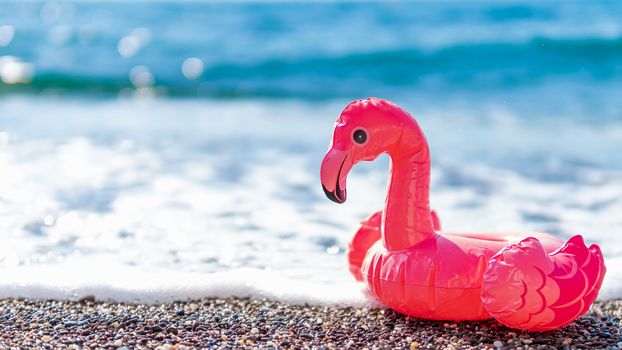 Inflatable pink flamingos on beach with white foam of sea wave. Summer, vacation, swimming, beach resort concept with pink flamingo. Banner. Copy space for text or design.