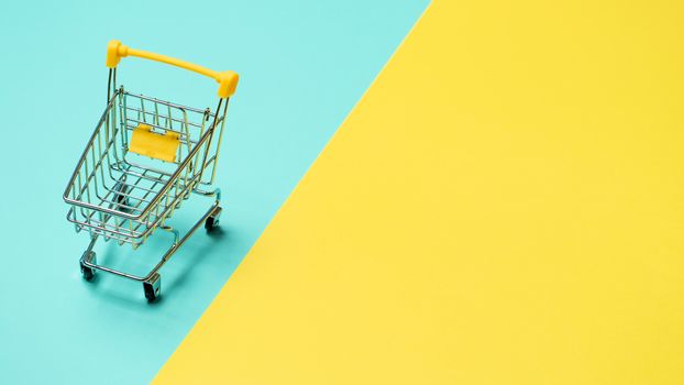 Empty miniature shopping cart on blue and yellow background. Toy trolley on bright colorful background, copy space for text or design.