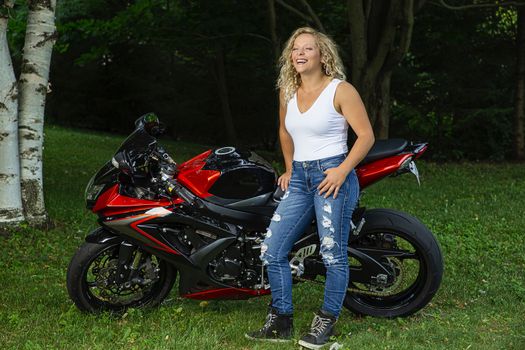 twenty something blond woman, wearing a pair of rip jean and white tank top, standing in front of a sport motocycle