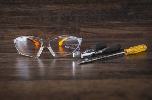 protective glasses, pliers and a screw driver against a dark wood background