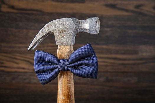 Old hammer with wood handle, wearing a blue bow tie, against a dark wood background