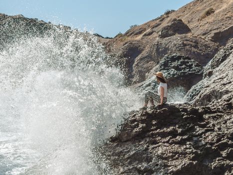 Woman sitting on cliff's edge near sea with big splashes.