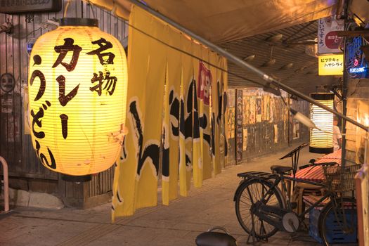 Night view of a traditional retro yellow rice paper lantern where it is written "Meibutsu Kare Udon" which means "Curry noddles Specialty" on underpass Yurakucho Concourse under the railway line of the station Yurakucho. Japanese noodle stalls and sake bars revive the nostalgic years of Showa air with old posters and placards glued to the walls of the tunnel.