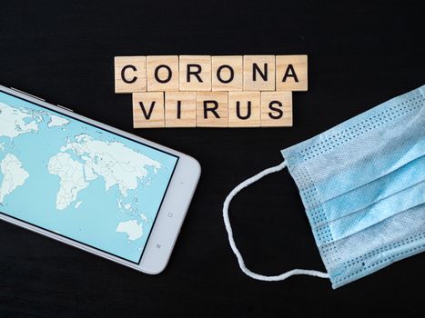 Coronavirus word made of wood block, world map on smartphone screen and breathing mask. Coronavirus text on dramatic atmosphere black wooden table. Coronavirus concept top view.